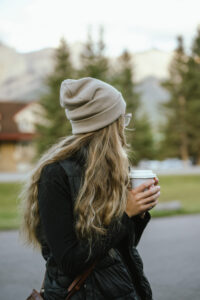 Woman in a beanie holding a cup, walking in nature with mountains and trees in the background during autumn.